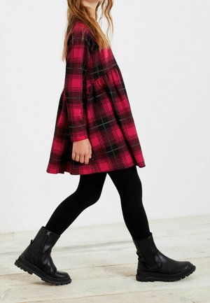 Robe à carreaux rose et noir avec des manches longues, une jupe évasée et une taille froncée. Associée à des leggings noirs et des bottes noires épaisses.