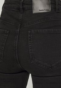 Jeans de ganga pretos com uma cintura alta, apresentando dois bolsos traseiros e uma etiqueta de marca na cintura. Tecido liso e texturizado.