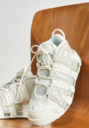 Paire de baskets blanches Nike Air More Uptempo avec un grand lettrage « AIR », posées sur une chaise en bois.
