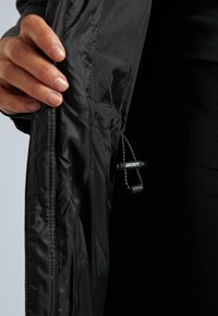 Melna jaka ar gludu, spīdīgu audumu. Iezīmē redzamu sašaurinājuma auklu un kabatu ar rāvējslēdzēju. Minimālisma dizains, nav redzamu rakstu.