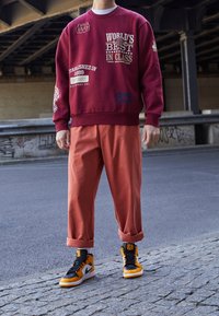 Felpa bordeaux con scritte e grafiche bianche, pantaloni arancioni comodi e sneakers alte nero-arancioni con dettagli bianchi.