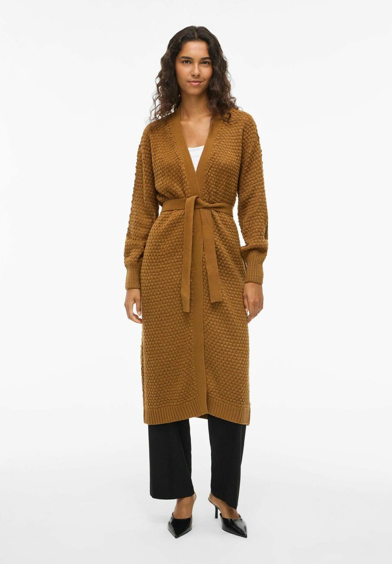 Cardigan en maille marron avec un motif texturé, devant ouvert, ceinture à la taille, manches légèrement surdimensionnées et poignets en côtes, associé à un pantalon noir.
