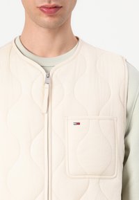 Chaleco acolchado beige con cierre de cremallera, presenta un bolsillo frontal y un sutil patrón de ondas. La tela parece suave con un diseño liviano.