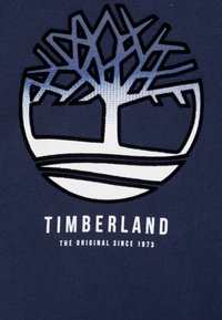 Mörkblå tyg har en central logotyp med en stiliserad trädesign i gradient blått och vitt, och fet, vit text nedanför som säger "TIMBERLAND."