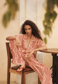 Roter und weißer gestreifter Jumpsuit mit Kragen, featuring wellenförmige Linien und offener Halslinie, kombiniert mit roten Sandalen, sitzend auf einem Holzstuhl.