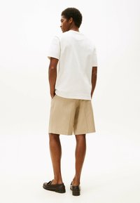 Camiseta blanca de algodón, pantalones cortos a rayas beige y mocasines negros. Modelo visto desde atrás, de pie sobre un fondo blanco.