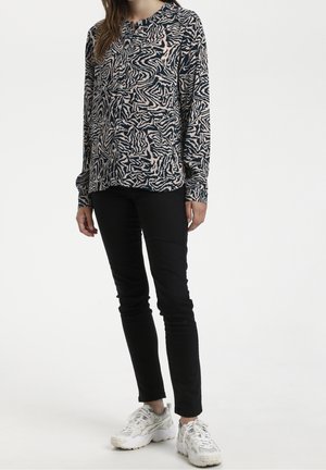 Vrouw met een blouse met lange mouwen in zwart en beige met een abstract patroon, zwarte nauwsluitende broek en witte bulk sneakers, staand tegen een witte achtergrond.