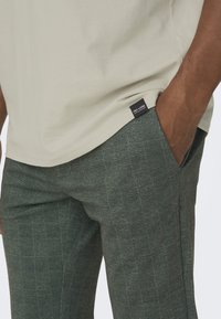 Lichtbeige t-shirt met een ronde hals en een logo-tag, gecombineerd met donkergroene geruite broeken met een zachte textuur.