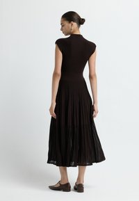 Tmavě hnědress, žebrované, bez rukávů, s upnutým vrškem a splývavou plisovanou sukní. Texturovaná tkanina a délka do poloviny lýtek, nošena s plochými botami.