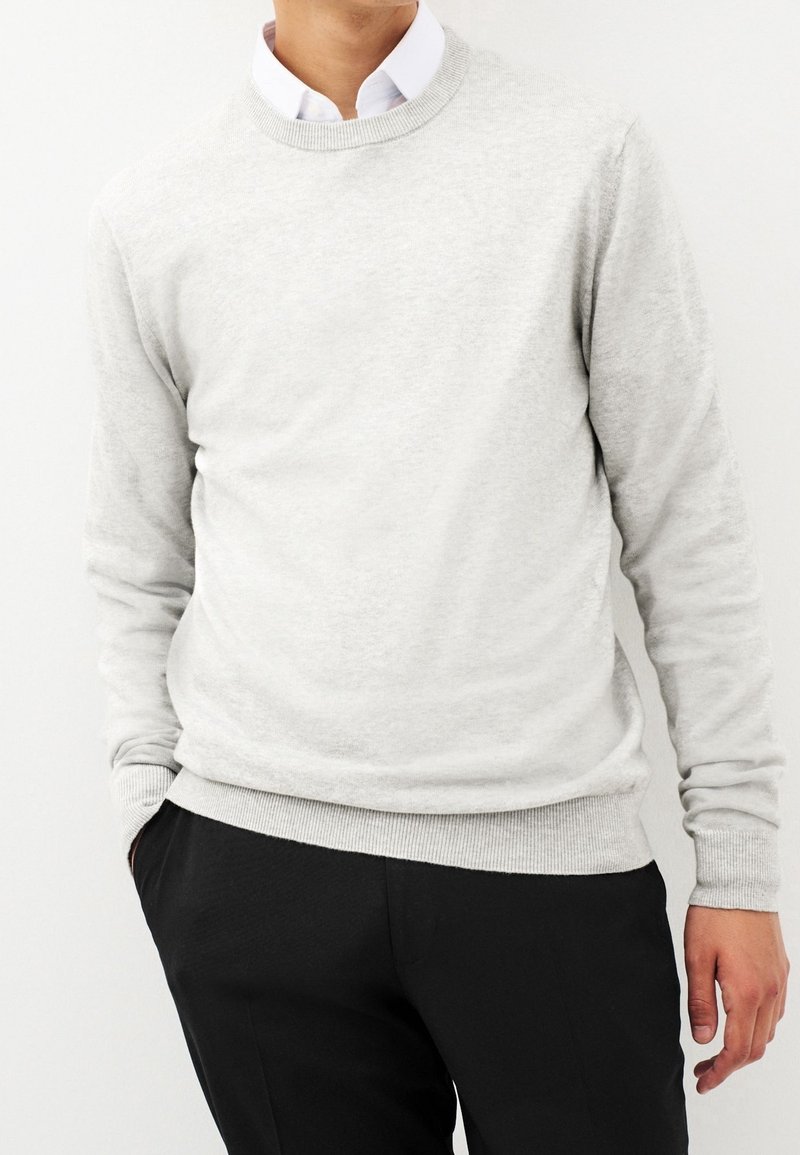 Homme portant un pull gris clair à col rond par-dessus une chemise blanche à col et un pantalon noir, debout avec une main dans la poche sur un fond blanc.