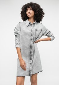 Szara sukienka koszulowa z denimu, z wyraźnym kołnierzykiem, zapięciem na guziki i pojedynczą kieszenią na piersi. Charakteryzuje się podwiniętymi rękawami i strzępionymi brzegami.