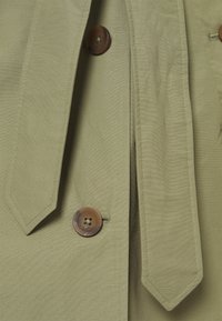 Khaki jas met een gestrikt taille, grote bruine knop en contrasterende stiksels. Het materiaal is glad met een lichte glans.