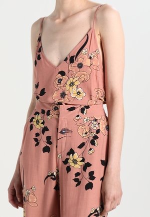 Kvinde iført en ærmeløs, lyserød buksedragt med blomsterprint, tynde stropper og V-hals mod en ensfarvet baggrund.