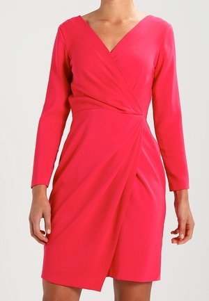 Kobieta ubrana w jaskraworóżową, długorękawową sukienkę kopertową z dekoltem w szpic, z marszczonym pasem i sięgającym do kolan dołem, na jednolitym tle.