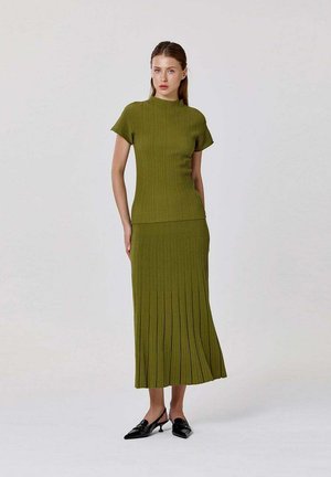 Femme debout portant un haut en tricot côtelé vert olive à manches courtes et une jupe midi plissée assortie avec des chaussures slingback noires à talons bas.