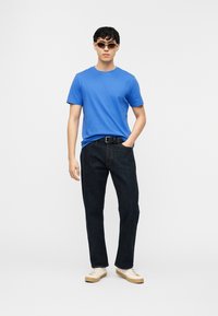 Άνδρας που φοράει γυαλιά ηλίου, μπλε κοντομάνικο t-shirt, σκούρο τζιν με μαύρη ζώνη και λευκά αθλητικά παπούτσια, στέκεται με το ένα του χέρι στην τσέπη.