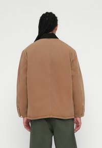 Brun bomuldsjakke med en sort fløjlskrage, synlige sømme og trykknapper. Designet har en afslappet pasform og oversized form.