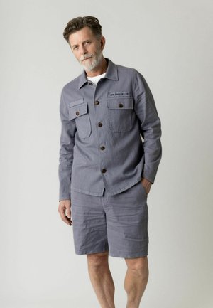 Mężczyzna w średnim wieku z siwymi włosami i brodą, ubrany w szarą kurtkę na guziki i dopasowane spodenki, z jedną ręką w kieszeni, stojący na jednolitym tle.