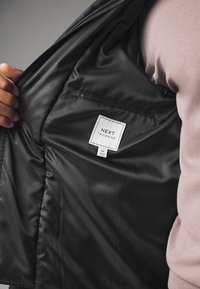 Zwarte jas binnenkant met een glad, glanzend voering, een klein zakje en een label met de tekst "NEXT TAILORING" en maat "M".