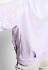 Světle fialová mikina s bílým nápisem "FREE STRIPS" na rukávu, žebrovaný lem a měkký bavlněný materiál s mírnou texturou.