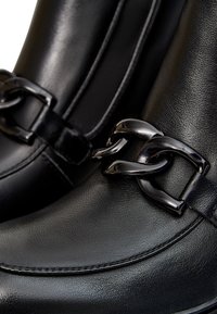 Botas de cuero negras con una textura suave, forma esbelta y un brillante acento de cadena negra en el lateral. Detalles de costura visibles.
