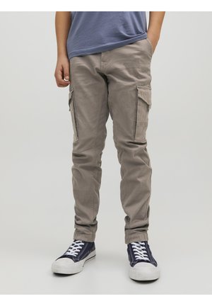 Person iført beige cargobukser, blå t-shirt og mørkeblå sneakers med hvide snørebånd, står med den ene hånd i lommen.