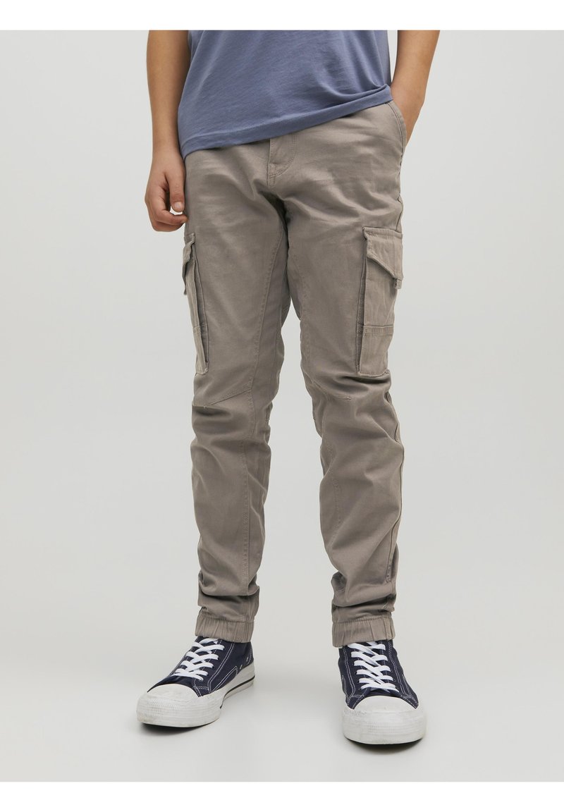 Personne portant un pantalon cargo beige, un t-shirt bleu et des baskets bleu marine avec des lacets blancs, debout avec une main dans la poche.