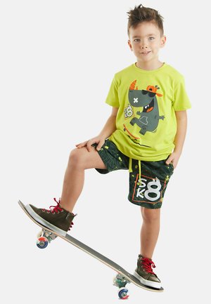 Leuchtend grünes T-Shirt mit verspieltem Motiv, gemusterte dunkle Shorts und schwarze Turnschuhe mit roten Schnürsenkeln. Kind steht auf einem Skateboard.