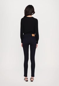 Kvinna med långt mörkt hår iklädd en svart långärmad tröja, mörka skinny jeans med en brun lapp och svarta sandaler, stående med ryggen mot kameran.