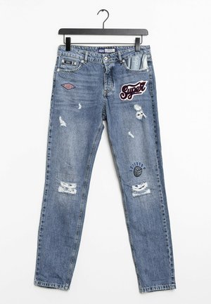 Niebieskie znoszone jeansy z łatami, w tym z napisem "Super", grafiką siatkówki i wzorzystą kieszenią, wiszące na czarnym wieszaku na białym tle.