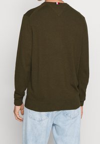 Oliwkowo-zielony sweter o fakturowanej powierzchni, z długimi rękawami i szwem w kształcie litery V na tyle dekoltu, zestawiony z jasnoniebieskimi dżinsami.