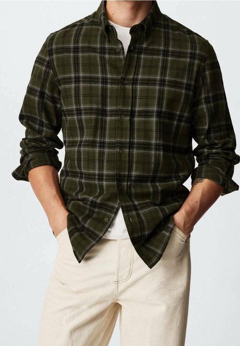 Homme portant une chemise à carreaux vert foncé boutonnée sur un sous-vêtement blanc avec les manches retroussées et un pantalon beige clair, mains dans les poches.
