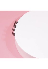 Black false eyelashes displayed on a round white surface against a pink background, featuring thin, wispy strands and clustered design.