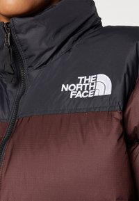 Veste isolante avec une partie supérieure noire et une partie inférieure bordeaux, col montant et logo blanc de marque sur la poitrine. Fermeture éclair à l'avant.