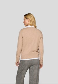 Beiger Pullover mit langen Ärmeln, Rundhals und geripptem Saum, über einem weißen Hemd getragen, kombiniert mit karierten Hosen. Minimale Details.