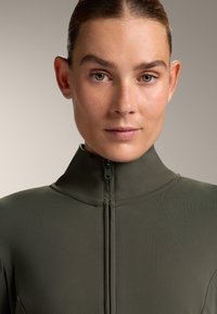 Close-up of a high-neck, zippered top in olive green fabric. The texture is smooth with a fitted design and subtle seams.