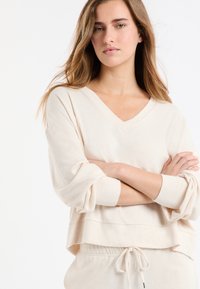 Kremowa, luźna bluza z dekoltem w serek, długimi, podwiniętymi rękawami i warstwowym dołem. Wykonana z miękkiej, teksturowanej tkaniny.