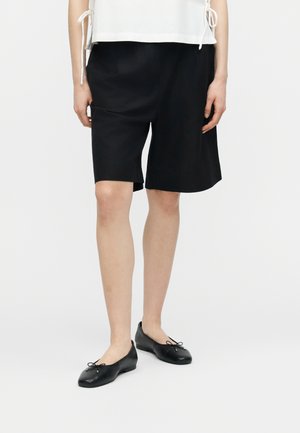 Persona con pantalones cortos negros hasta la rodilla, camiseta blanca con lazos laterales y bailarinas negras, de pie contra un fondo blanco liso.