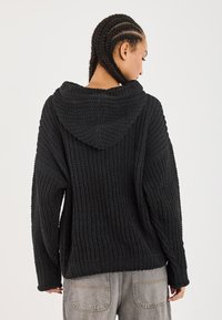 Zwarte gebreide hoodie met een brede, gestructureerde design en ruime pasvorm. Bevat een grote capuchon en ribgebreide manchetten en onderrand.