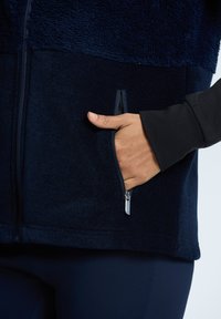 Marineblaue Fleecejacke mit strukturiertem oberen Bereich, glattem unteren Abschnitt und seitlichem Reißverschlusstasche. Mit schwarzem Ärmel-Detail.