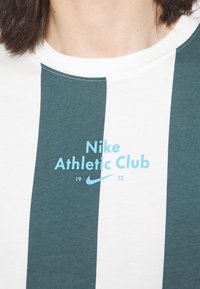 Bílý a zelený pruhovaný tričko vyrobené z bavlny, s grafikou "Nike Athletic Club" v světle modré barvě a malým logem Nike. Jednoduchý design s kulatým výstřihem.