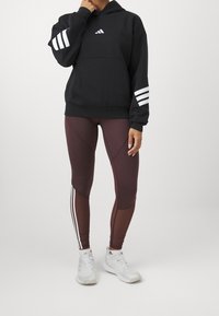Czarna bluza z kapturem z białymi paskami na rękawach, zestawiona z brązowymi legginsami z bocznymi paskami i fakturowymi panelami oraz białymi sneakersami.