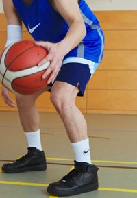 Sorte høyde basketballsko med hvite detaljer, iført blå ermeløs trøye og marineblå shorts, dribler en rød og hvit basketball.
