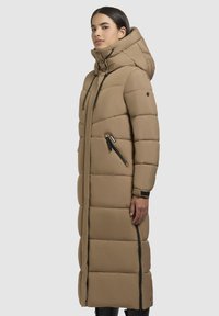 Cappotto lungo imbottito color beige, con colletto alto, grande cappuccio e tasche laterali con zip e accenti neri. Esterno liscio e resistente.