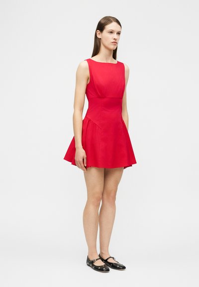 Γυναίκα που φοράει αμάνικο κόκκινο μίνι φόρεμα και μαύρα μπαλαρίνες, στέκεται μπροστά σε ένα ομοιόμορφο ανοιχτό γκρι φόντο, έχοντας ελαφρώς κλίση προς τα αριστερά.