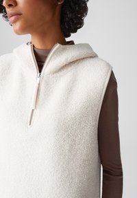 Cremfarbener Fleece-Weste mit Kapuze, ausgestattet mit einem silbernen Reißverschluss und Besatz. Die Weste wird über einem braunen Langarmshirt getragen, das eine strukturierte Oberfläche zeigt.