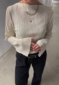 Krémový plisovaný top se širokými rukávy, doplněný černými kalhotami. Zlaté vrstvené náhrdelníky a prsteny dokreslují vzhled, zdůrazňují textury a barvy.