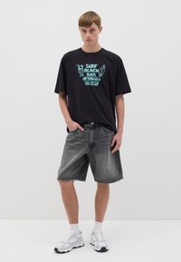 Camiseta negra con texto gráfico turquesa "SURF BEACH BAR" combinada con pantalones cortos de mezclilla grises y zapatillas blancas con acentos grises.