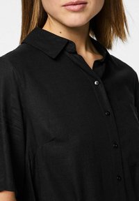 Juodas marškinėliai su mygtukais, pagaminti iš lygios, audinės medžiagos. Turi apykaklę, trumpas rankoves ir kišenę kairėje pusėje.