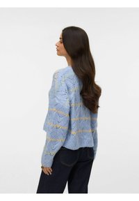 Hele sinine kootud kampsun, mis on kaunistatud pitsilise mustriga ja kollaste aktsentidega, ümar kaelus ja lühendatud alumine äär, kantud tumedate teksadega.